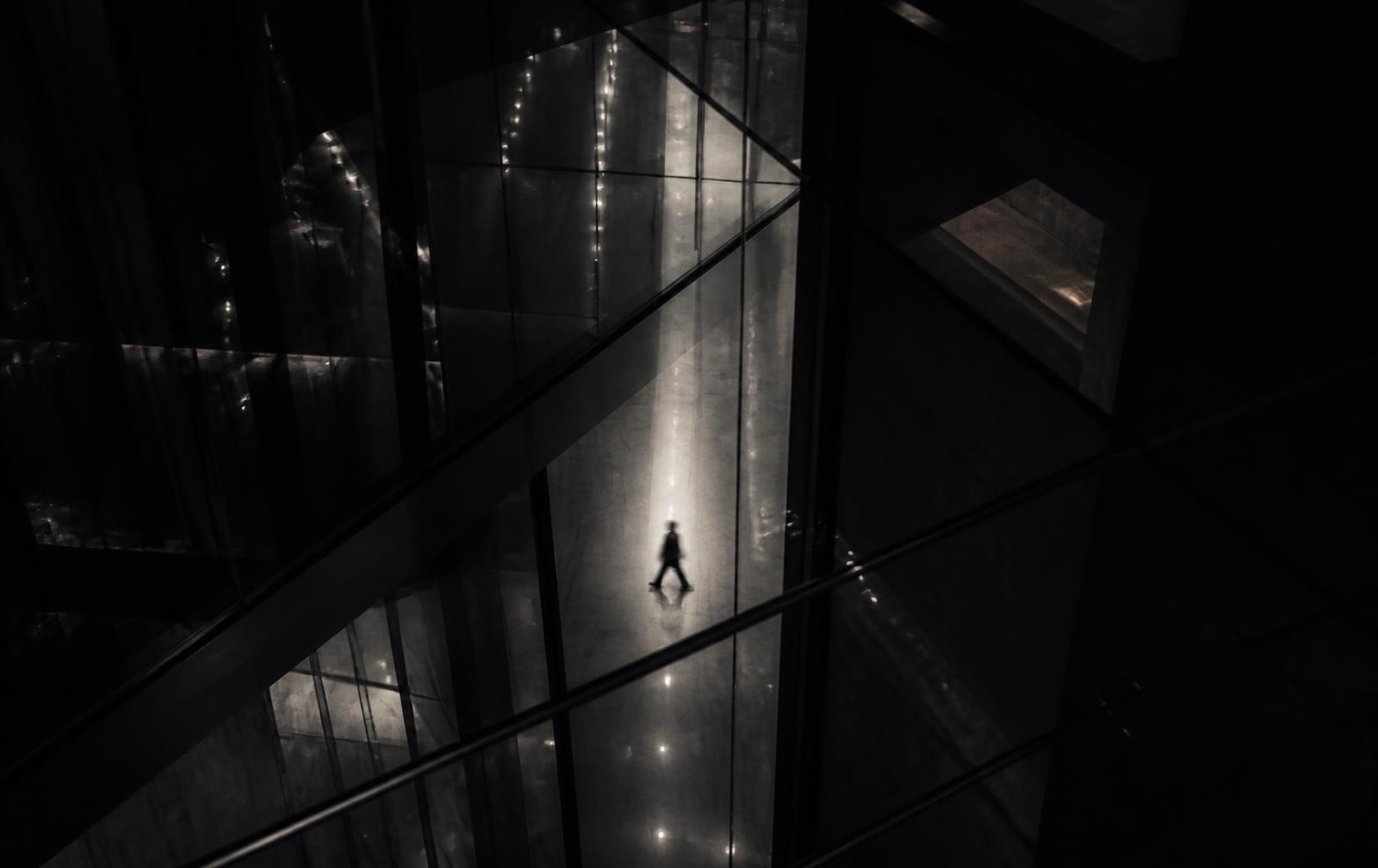 Dark glass atrium at night with a single distant silhouette crossing the floor